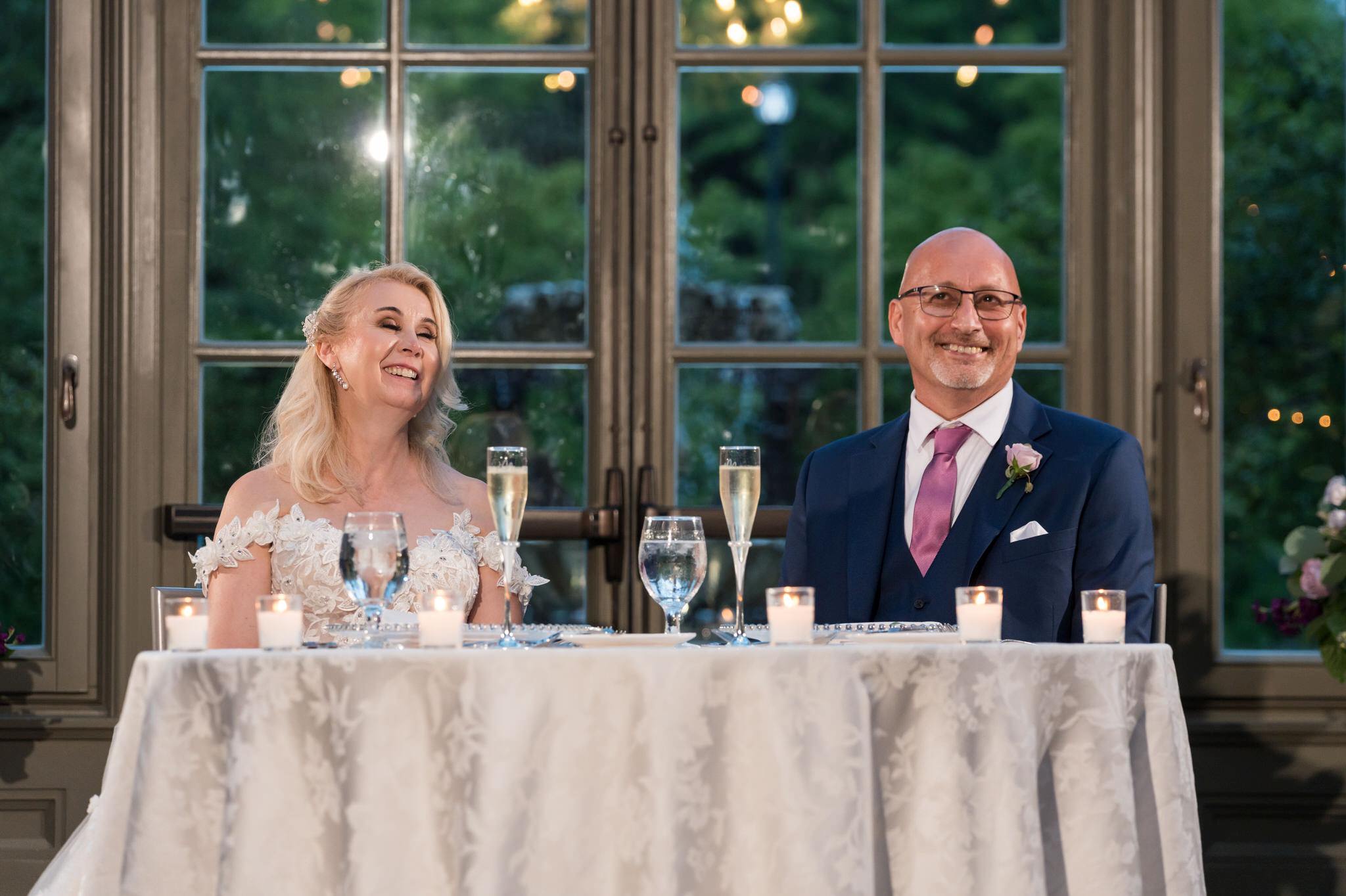 A bride and groom, sitting at a sweetheart table, laugh during their Royal Park wedding reception.  