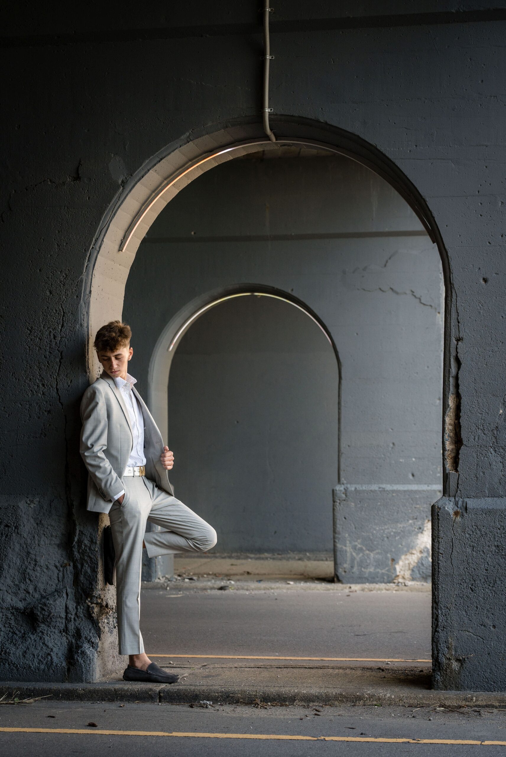 Male, wearing a gray suit, poses for a senior photo in Detroit against an archway.