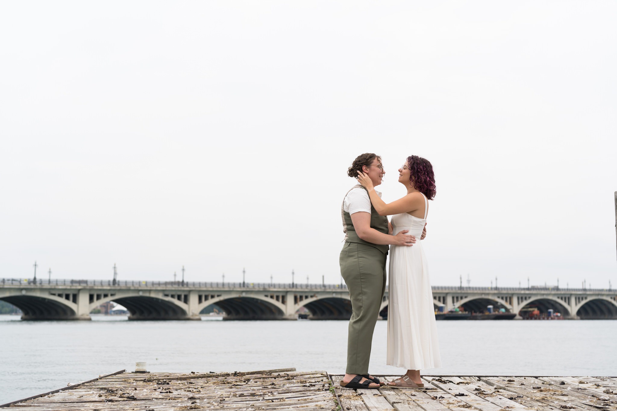 A same sex couple poses on a dock on their wedding day on Belle Isle with a bridge in the background.  