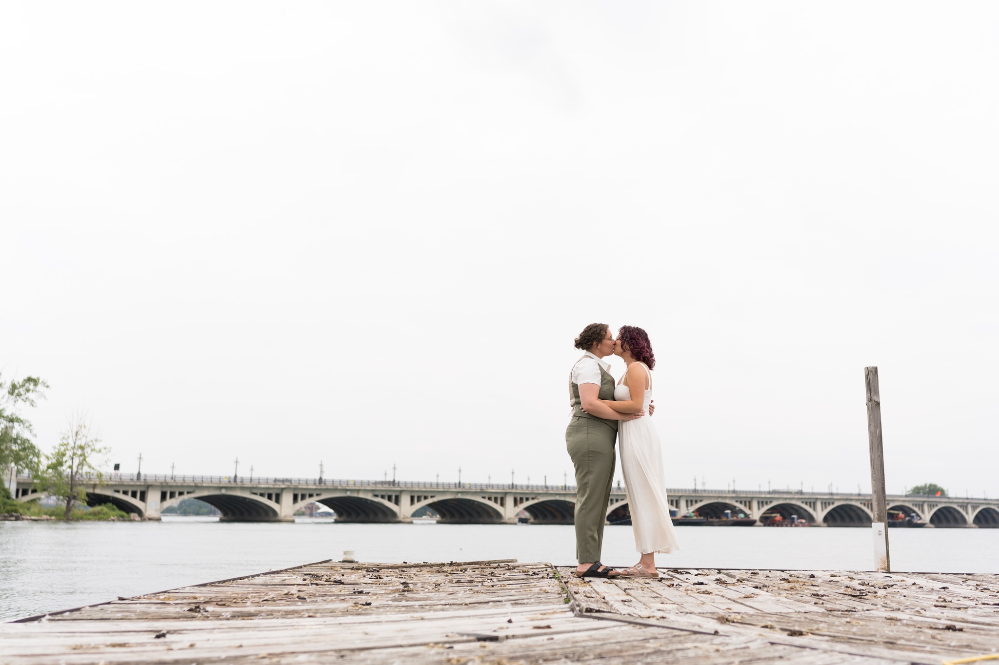 A same sex couple kisses on their wedding day on Belle Isle with a bridge in the background.  