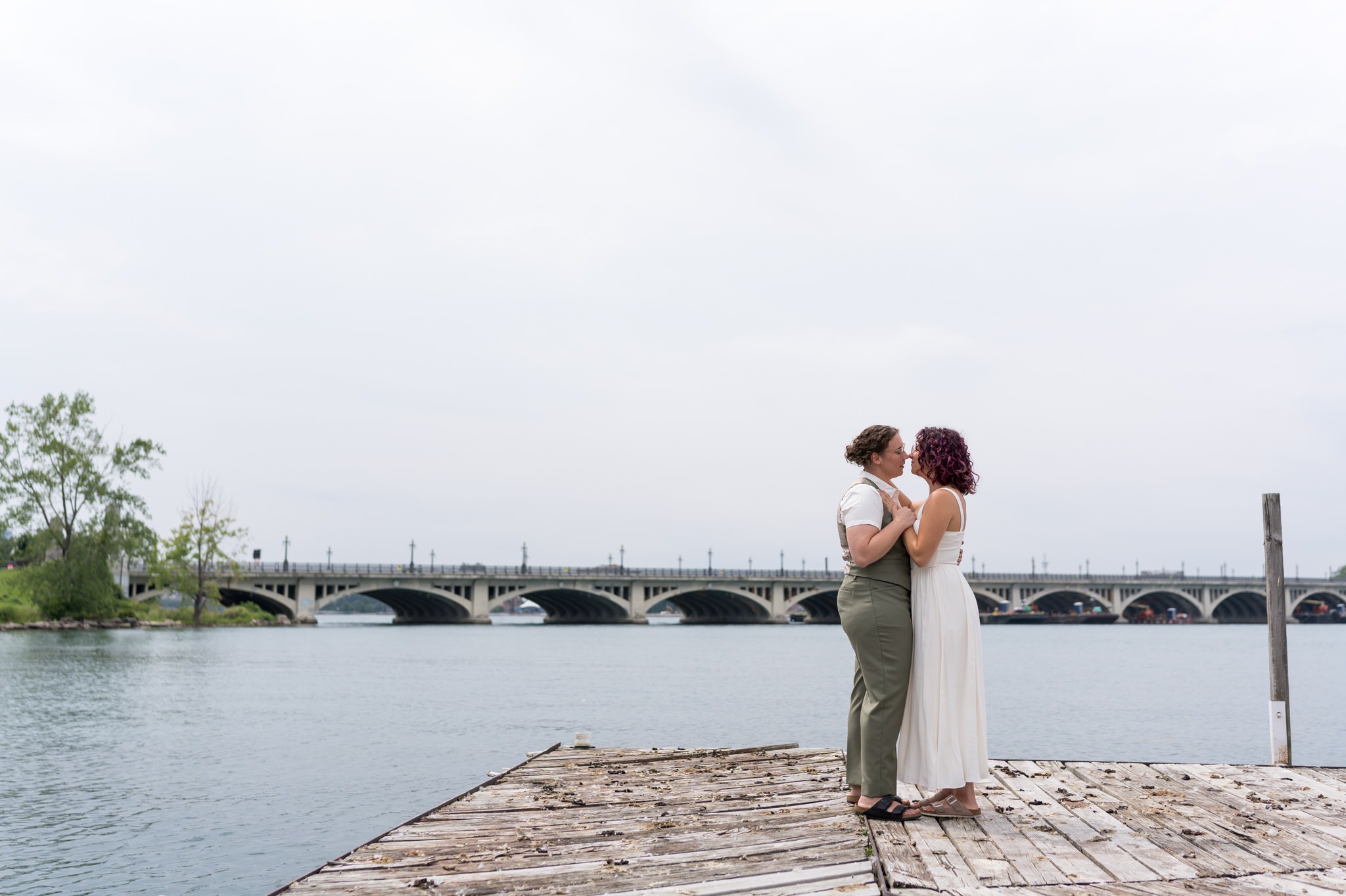 A same sex couple poses on their wedding day on Belle Isle with a bridge in the background.  
