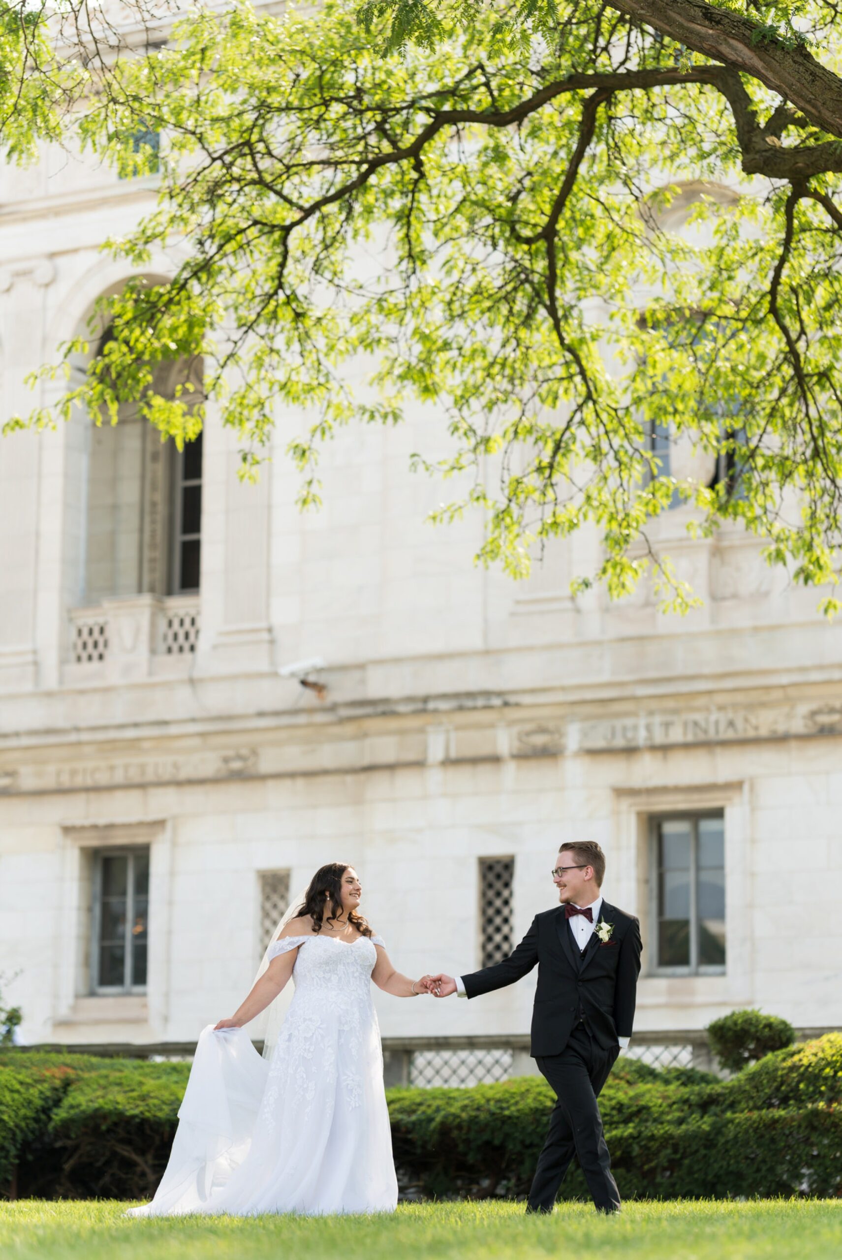 A bride and groom, holding hands, walk across the front lawn of the Detroit Public library on their wedding day. 