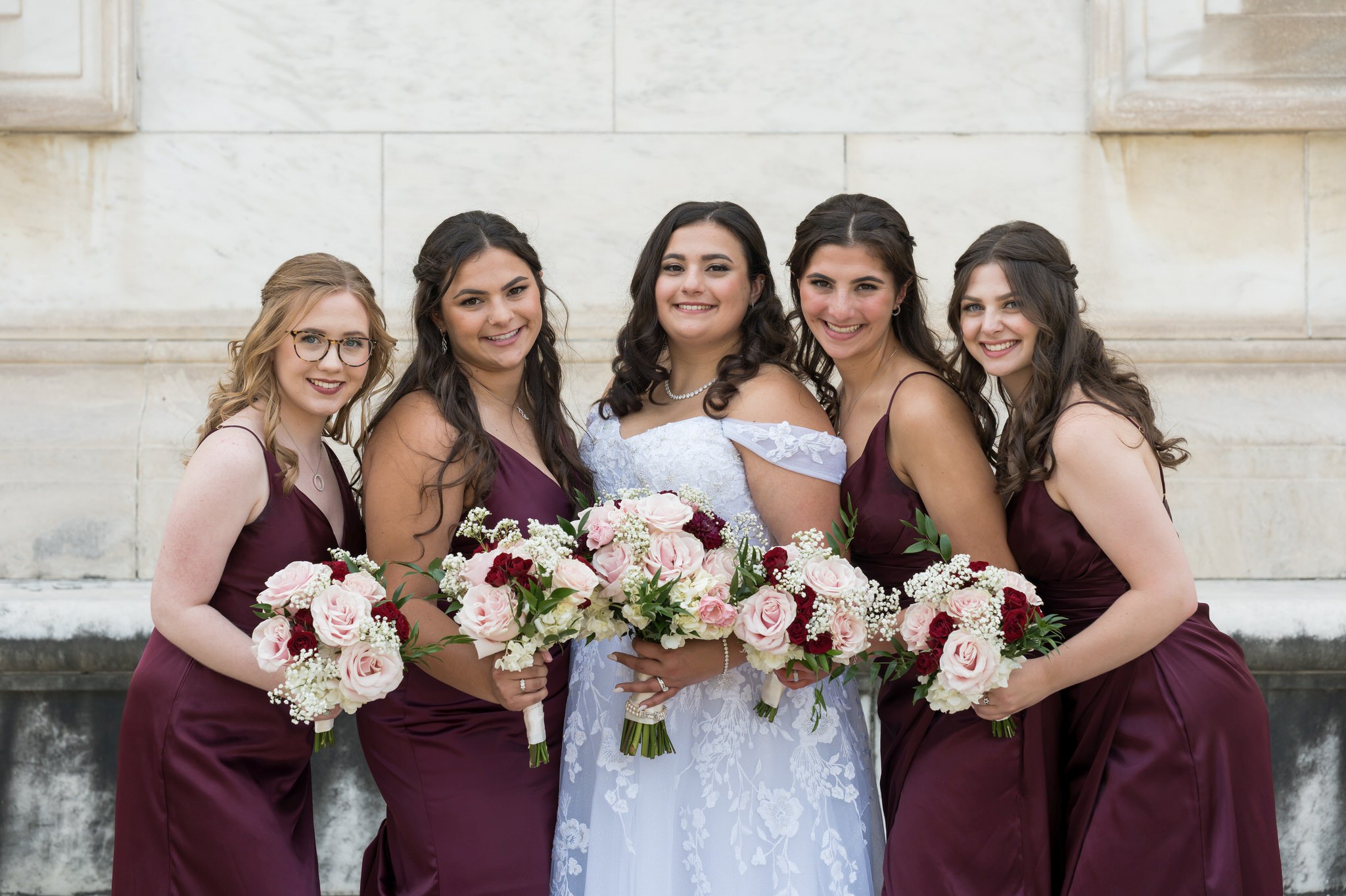 Bridesmaids, wearing maroon dresses, pose outside of the Detroit public library on a wedding day.  