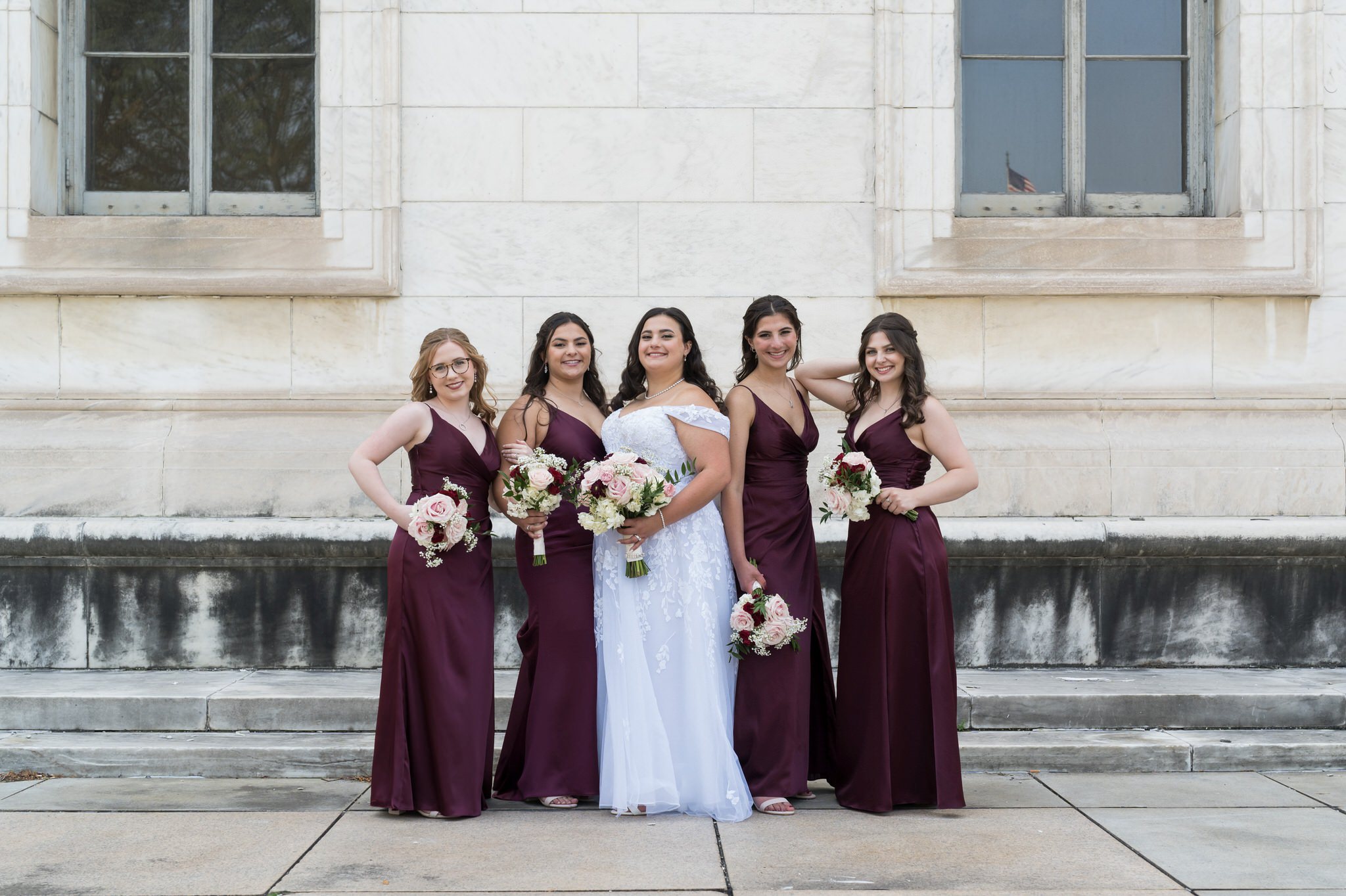 Bridesmaids, wearing maroon dresses, pose outside of the Detroit public library on a wedding day.  