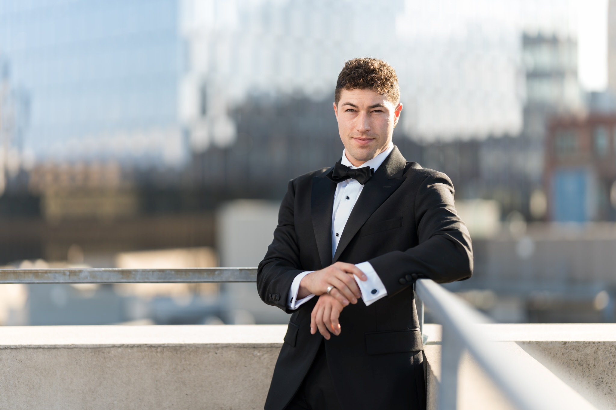 A groom poses, leaning on a railing, with a blurred out Detroit skyline in the background.  