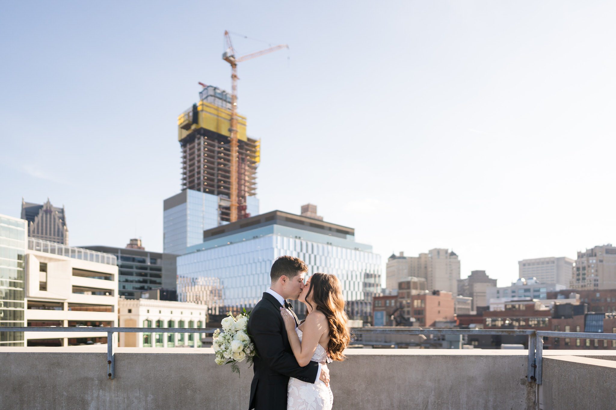 A bride and groom kiss on a Detroit rooftop with the skyline in the background.   