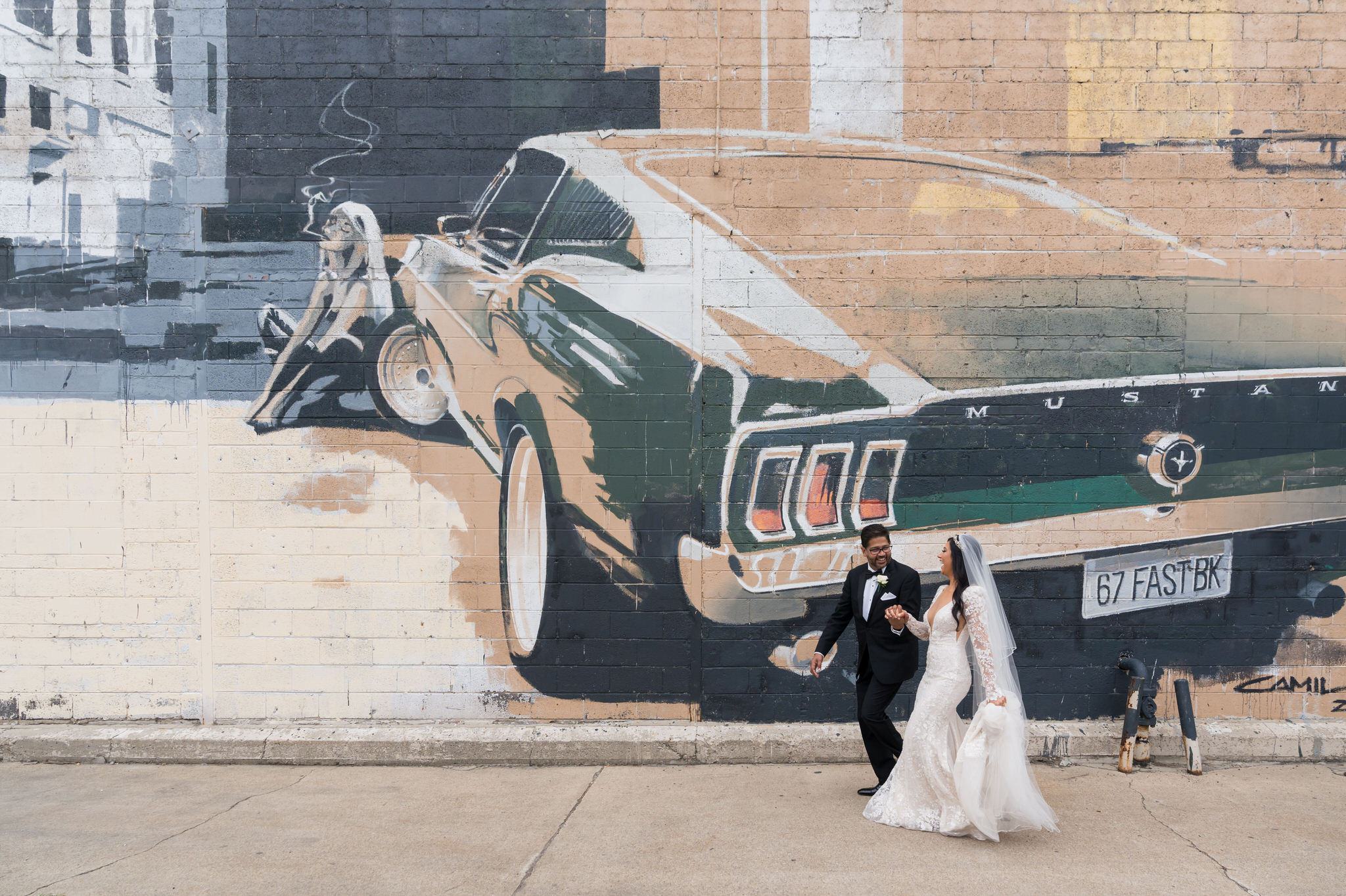 A bride and groom hold hands and run in front of a large mural on their wedding day in Eastern Market, Detroit. 