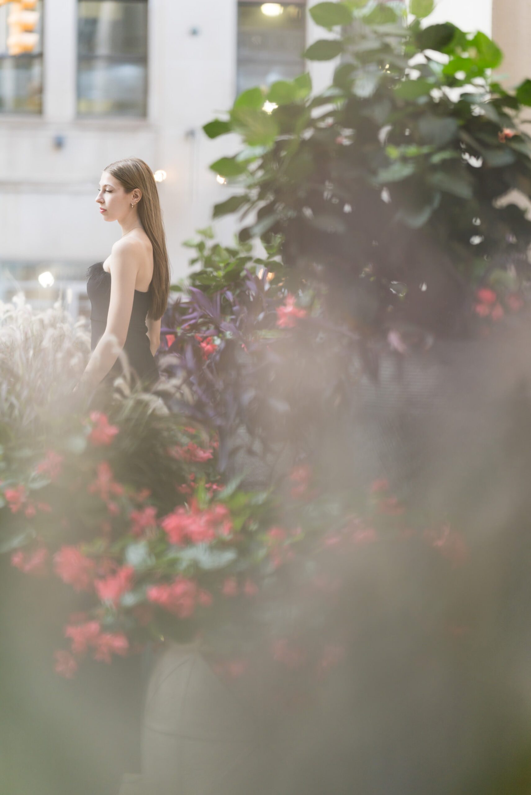 A woman wearing a black dress and pink heels poses among some greenery for her Detroit senior pictures.  