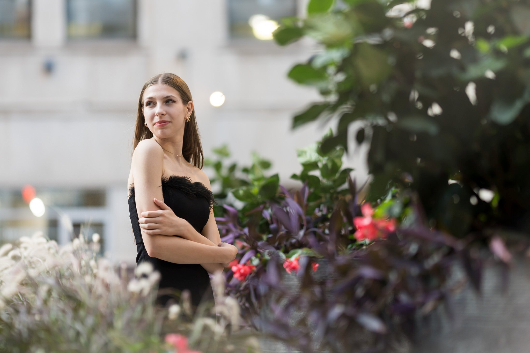 A woman wearing a black dress and pink heels poses among some plants for her Detroit senior pictures.  