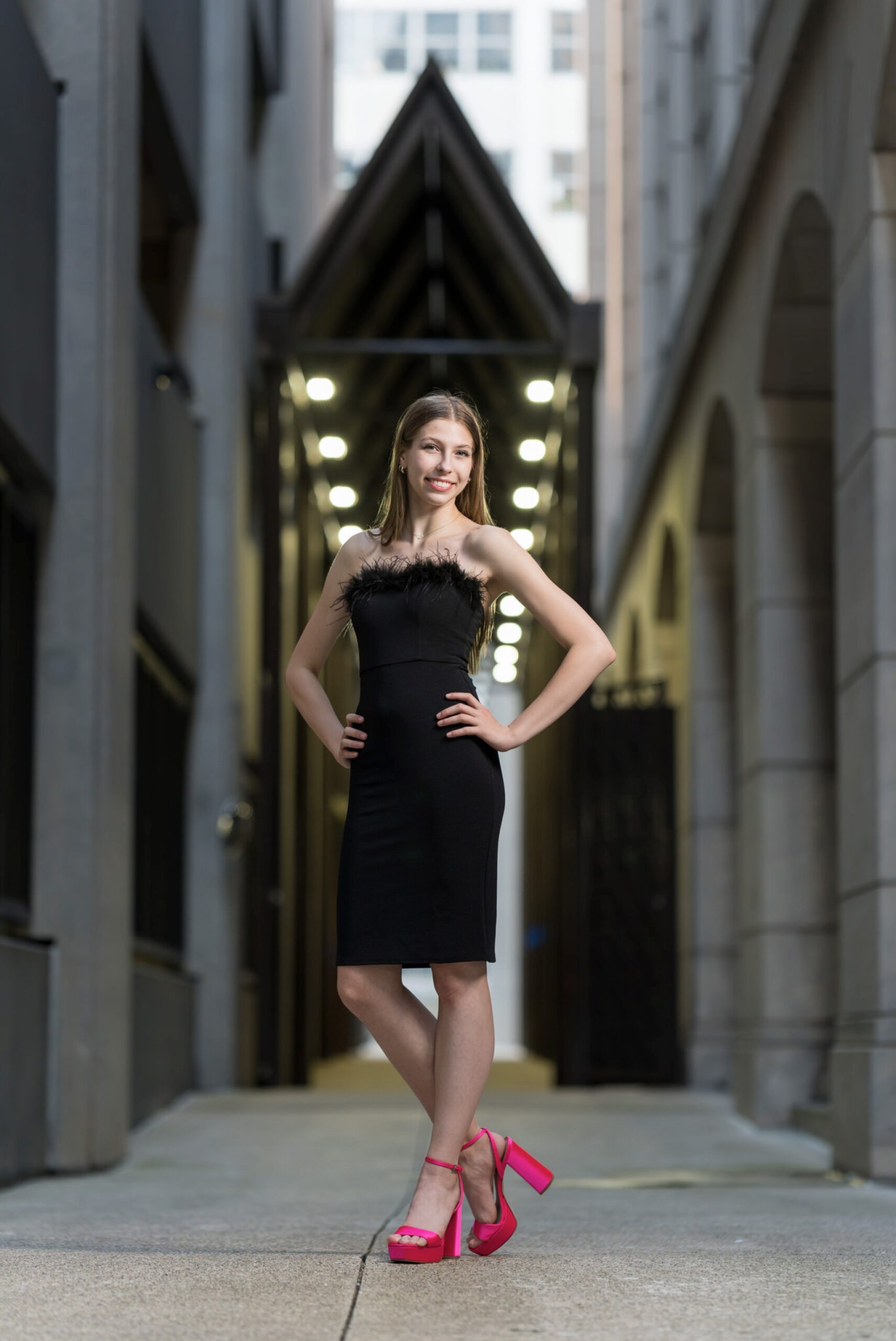 A woman wearing a black dress and pink heels poses in an alleyway for her Detroit senior pictures.  