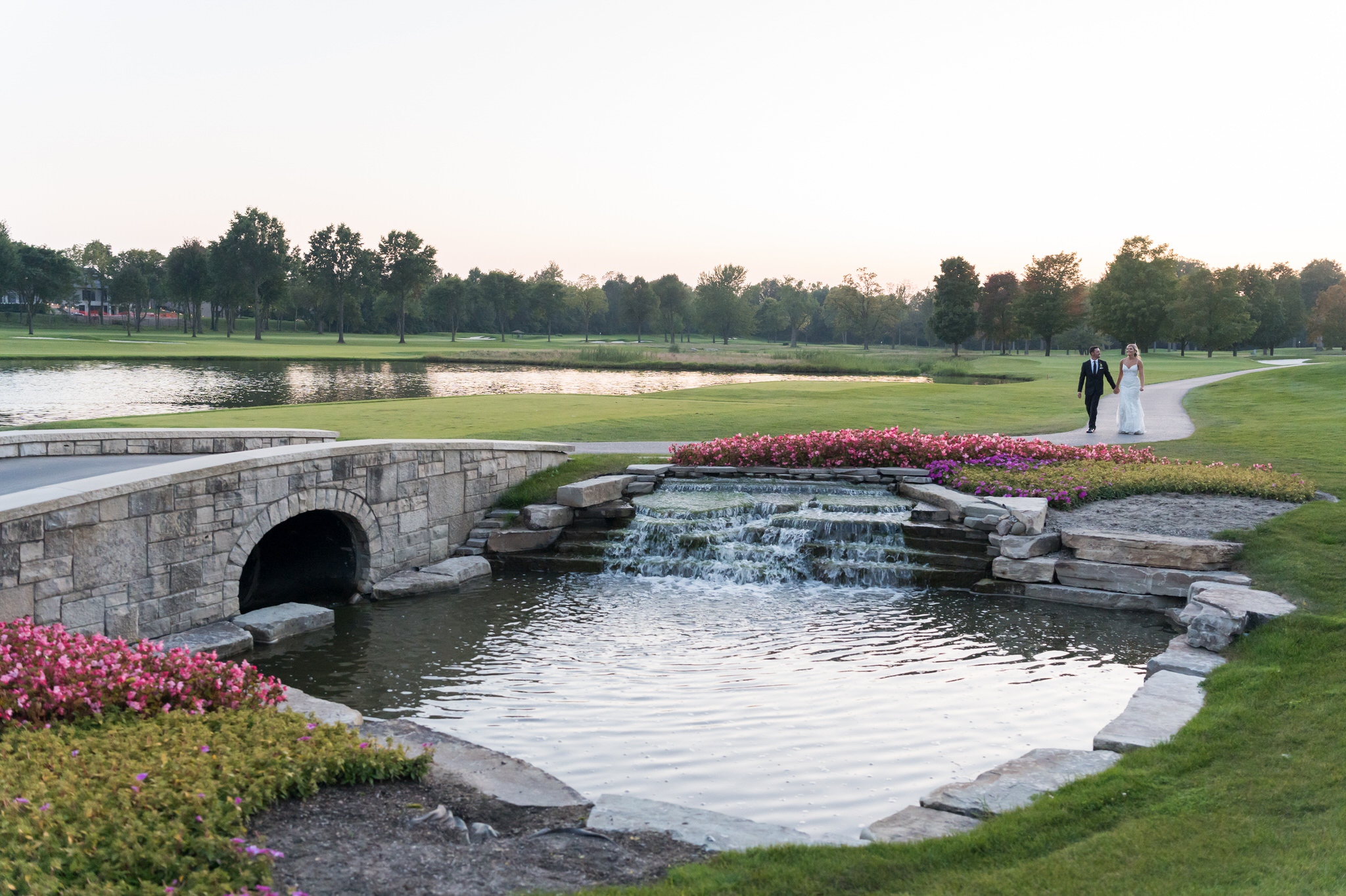 A bride and groom walk over the stone bridge at their Walnut Creek Golf Club wedding in South Lyon, MI.  