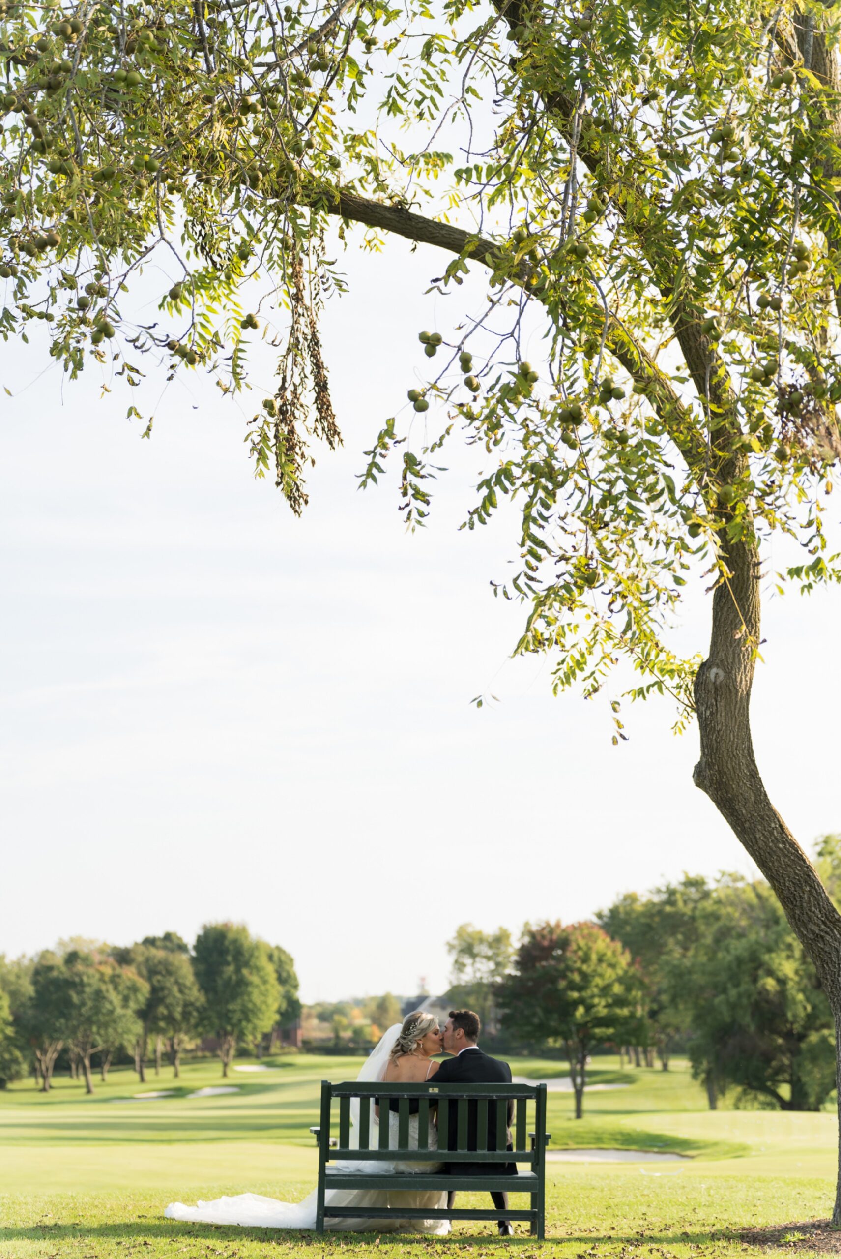 A bride and groom kiss while sitting on a green bench at their Walnut Creek Golf Club wedding in South Lyon, MI.  