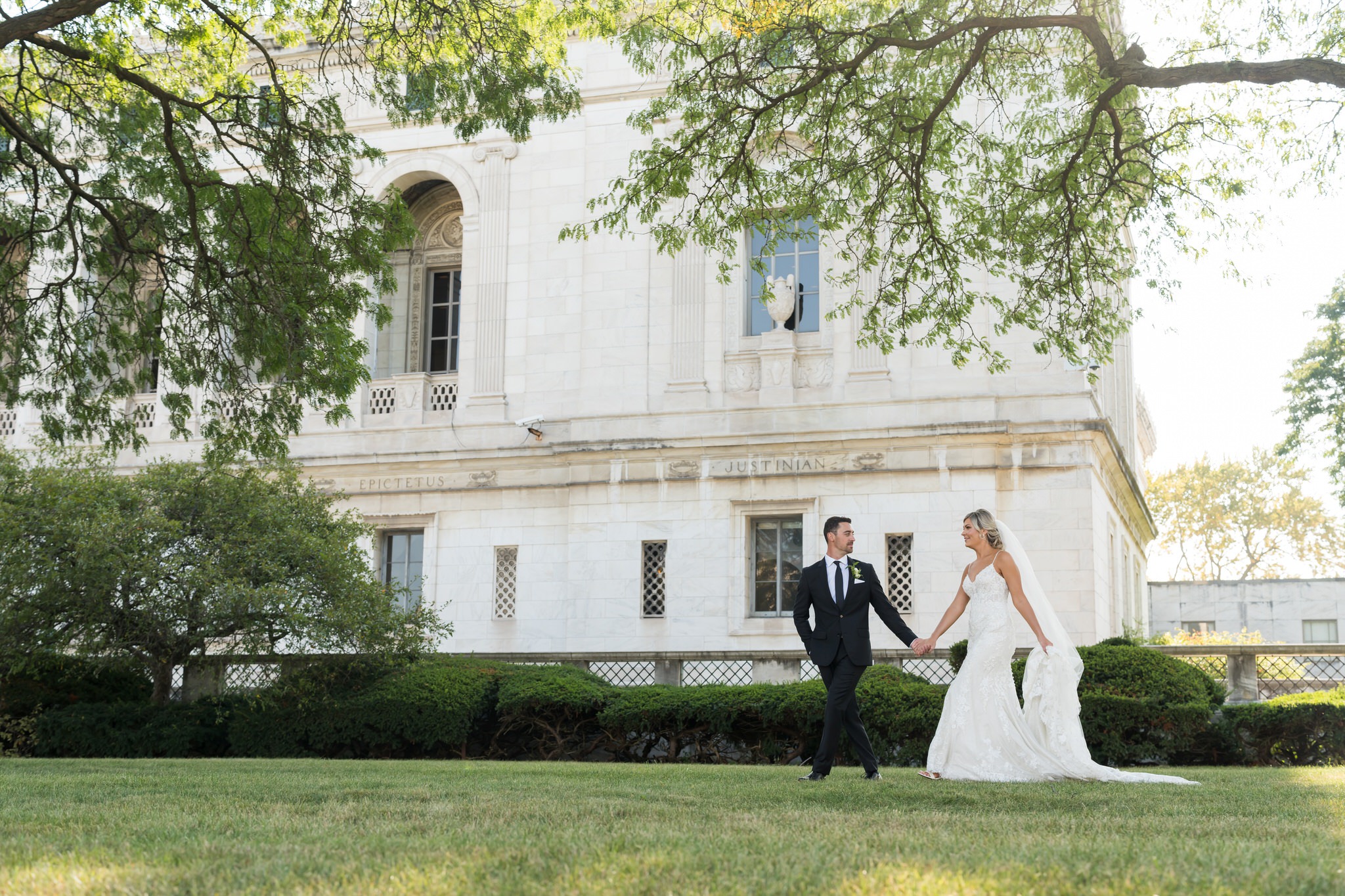 A bride and groom hold hands and walk across the grass at the Detroit Public Library on their wedding day.  