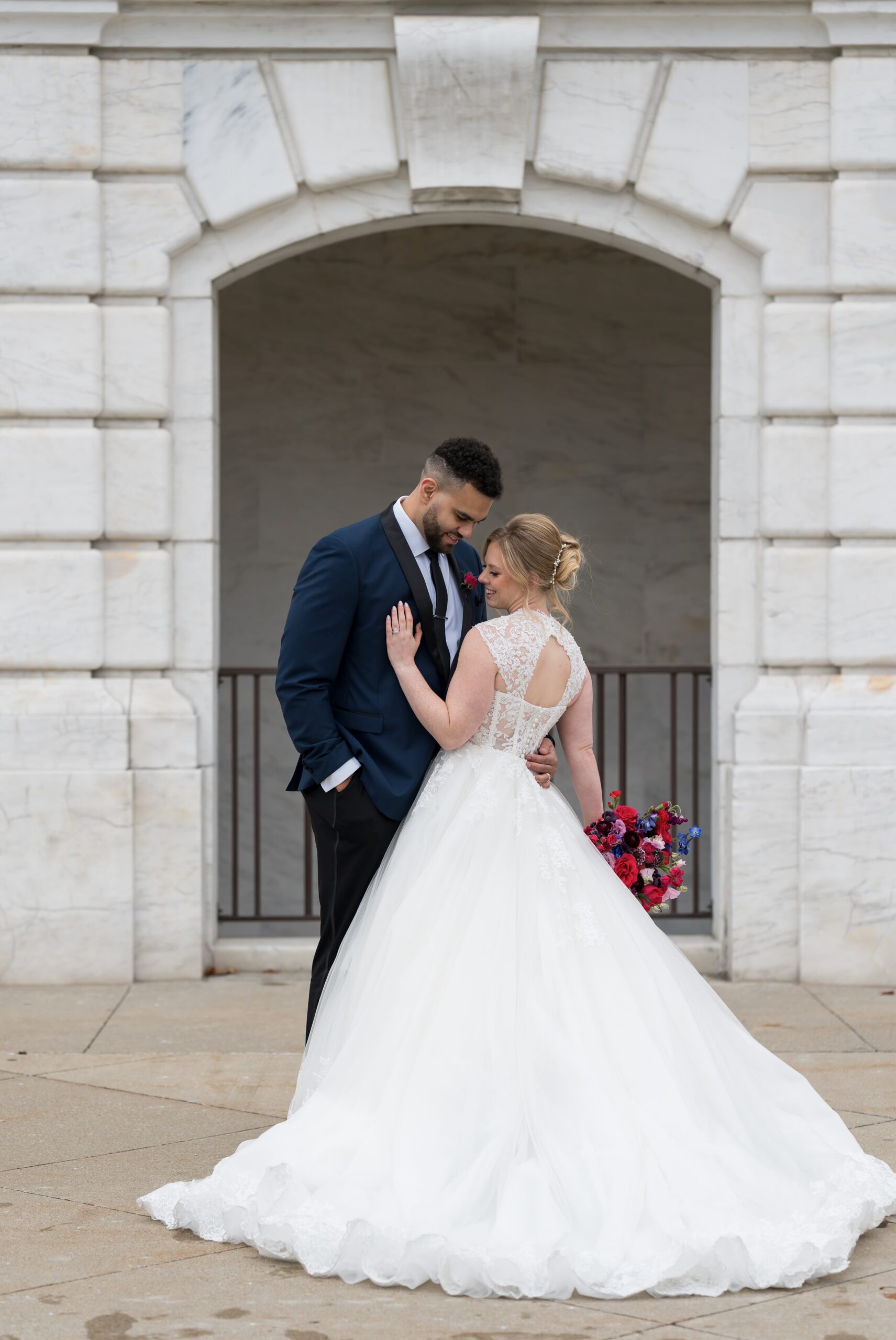 A couple, framed by a white marble arch behind them, touch foreheads on their wedding day.  