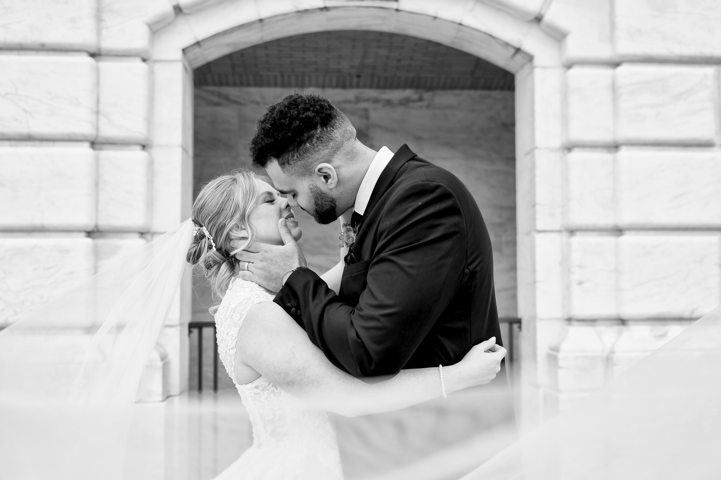 A couple, framed by a white marble arch behind them, almost kiss as her veil blows in the foreground.    