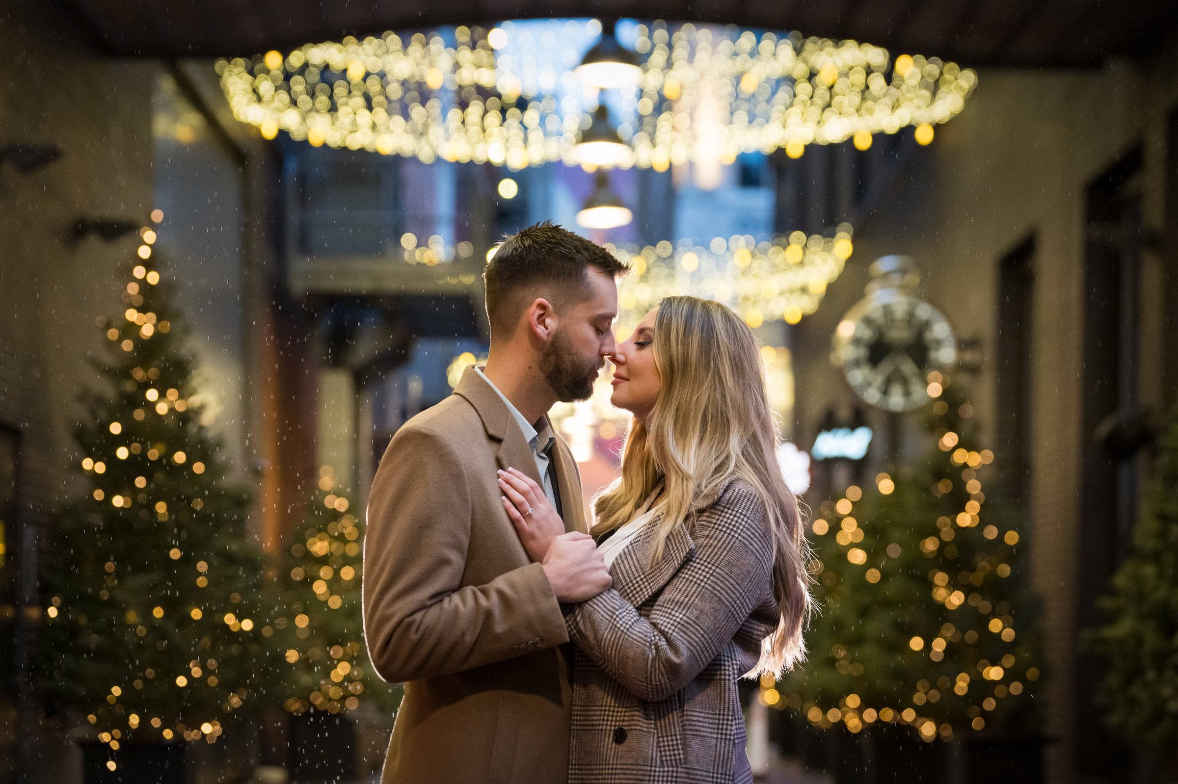 A couple, with Christmas trees in the background, pose under the twinkle lights of Parker's Alley after their proposal. 