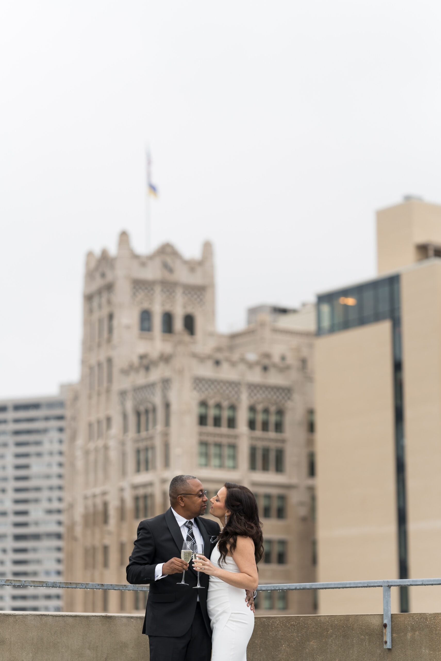 A bride and groom, holding champagne glasses, almost kiss with Detroit skyscrapers behind them on their wedding day.  