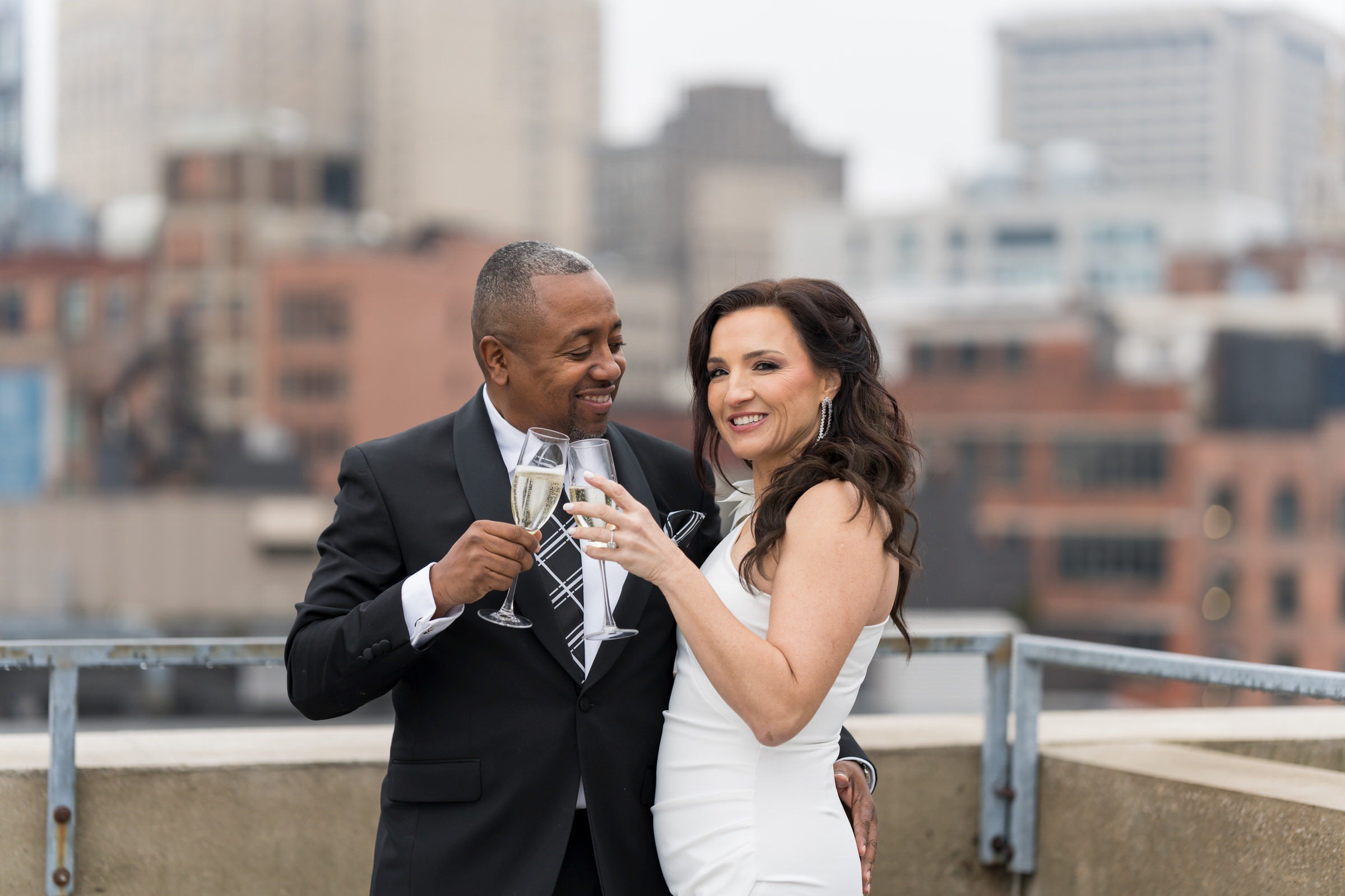 A bride and groom cheers champagne glasses with a city skyline behind them in Detroit on their wedding day.  