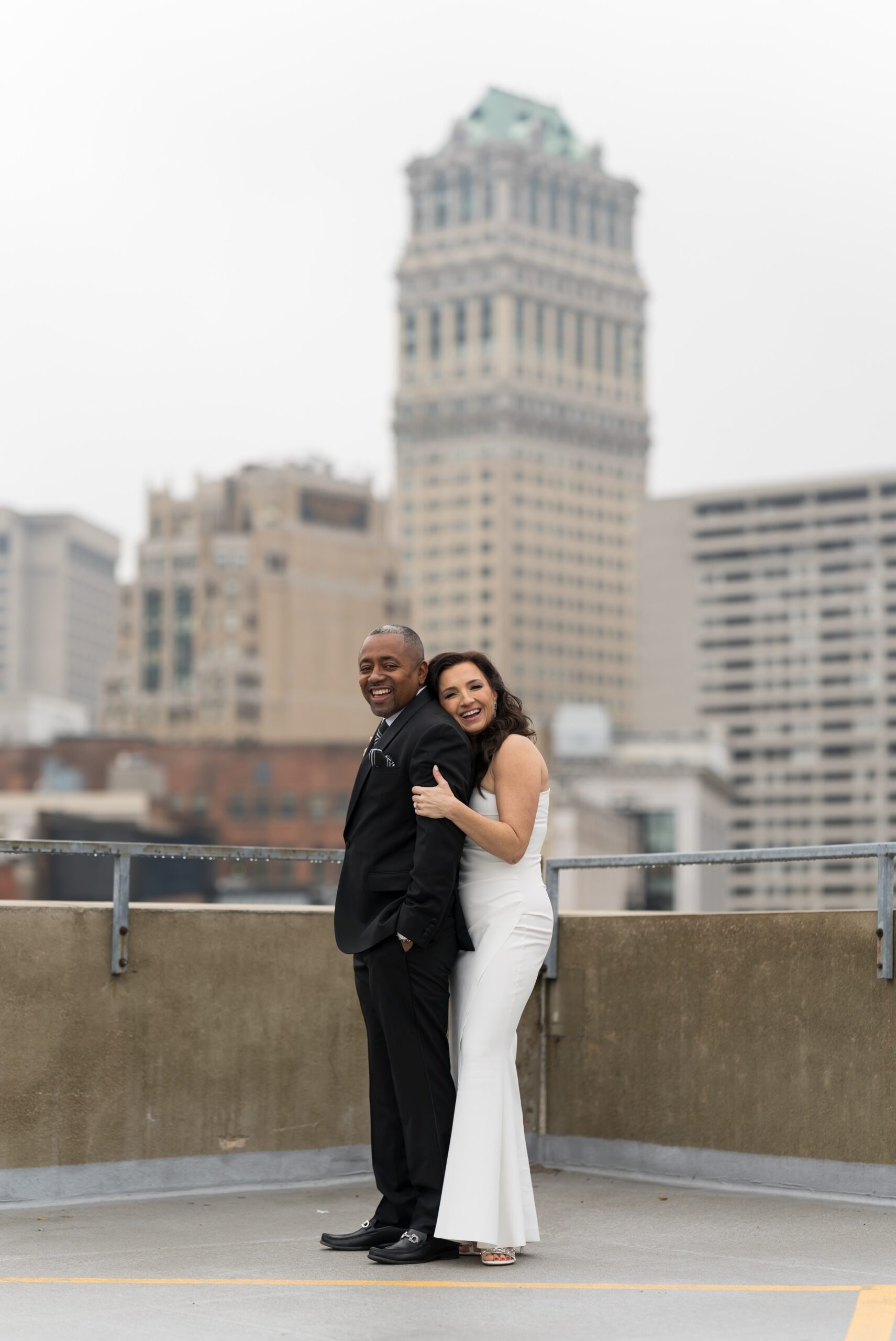 A bride hugs her groom from behind, laughing, atop the Detroit Opera House parking garage on their wedding day. 
