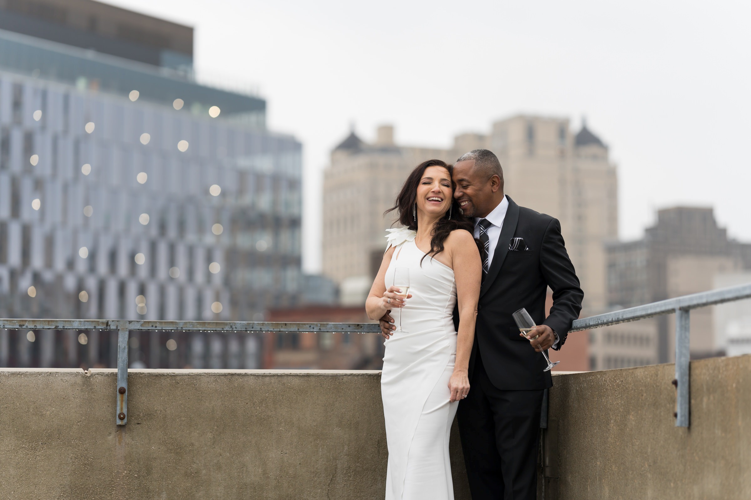 A couple, holding champagne glasses, laugh atop the Detroit Opera House parking garage on their wedding day. 