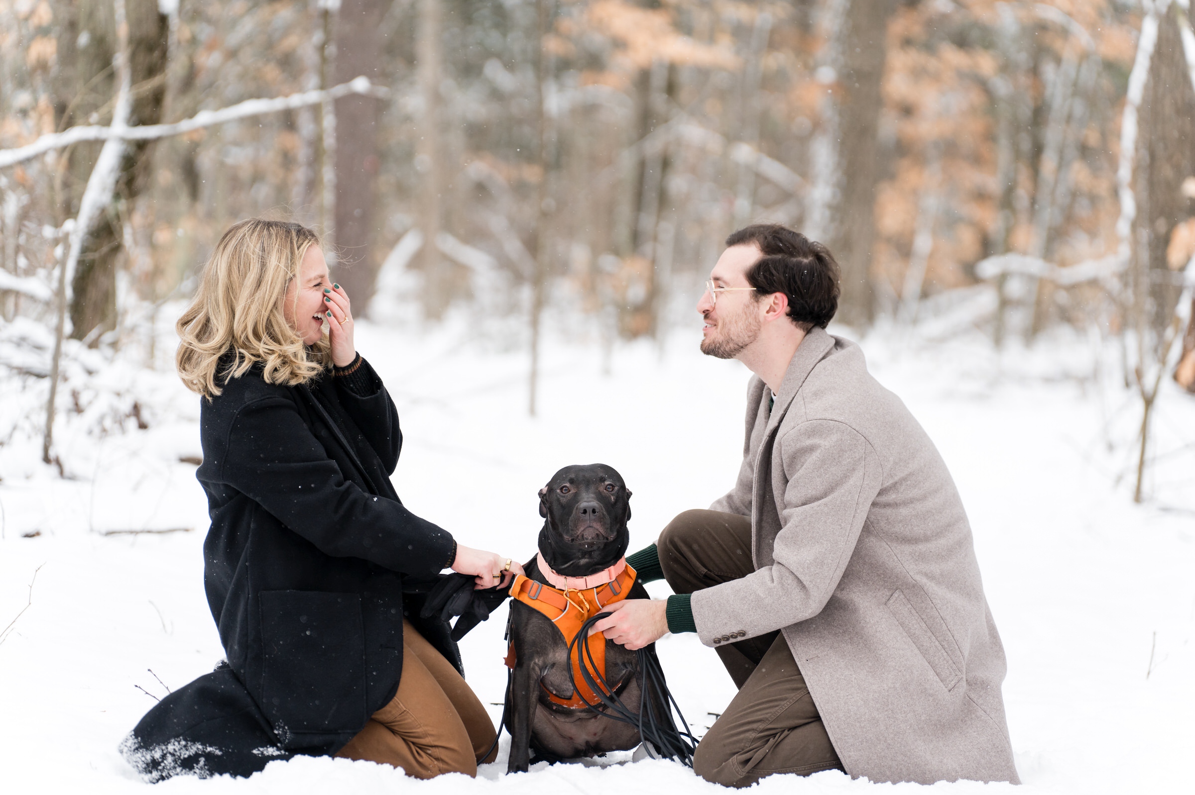 A couple, both kneeling in the snow, laugh and pet their dog after their Getaway proposal.  