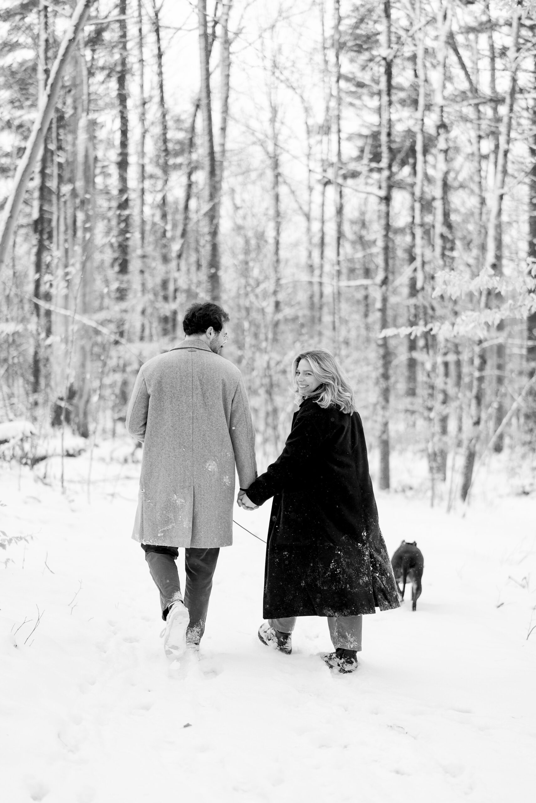 A couple, holding hands and walking a dog, walks along a snow covered path after their Getaway proposal.