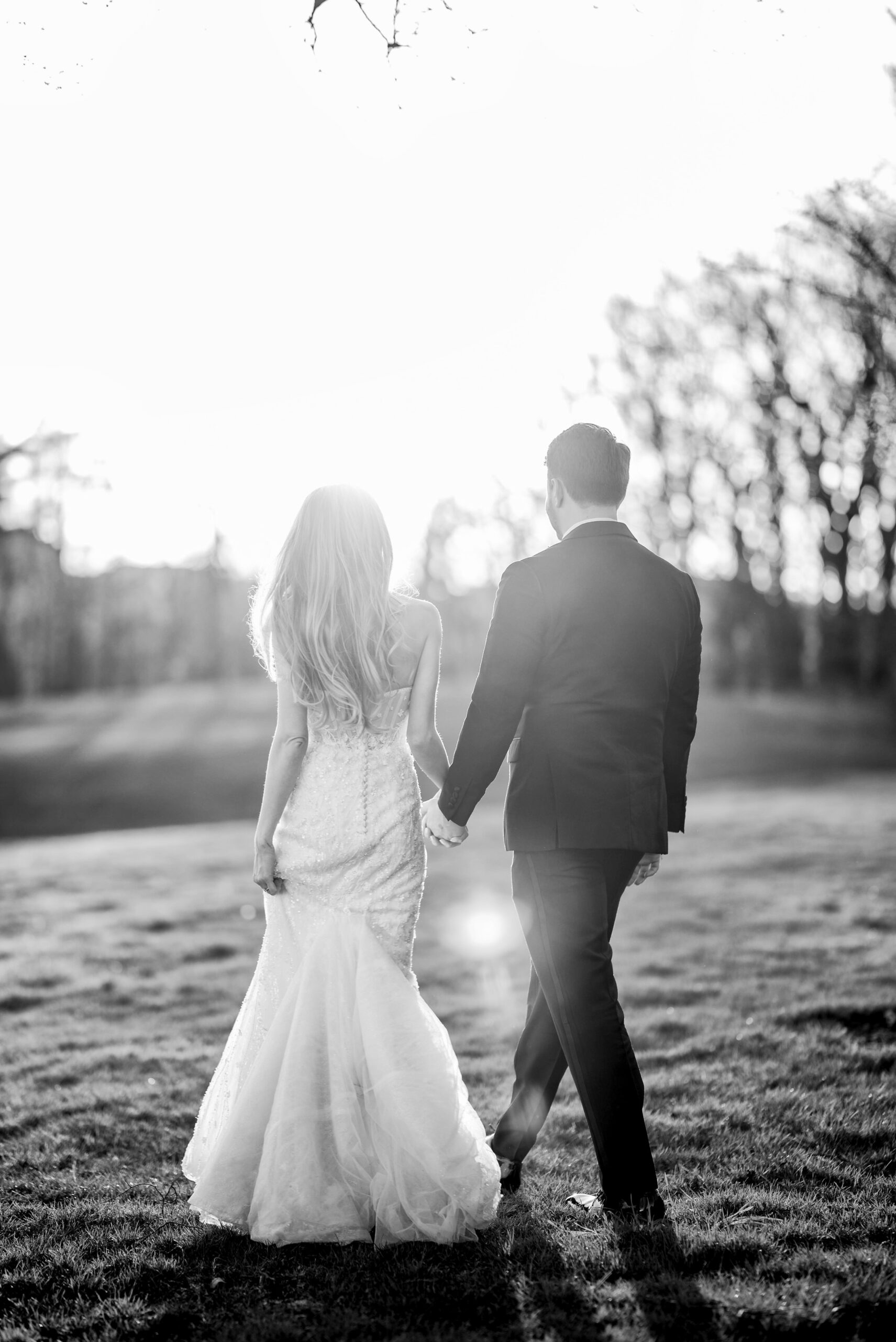 A couple, holding hands, walks away from the camera during a golden hour photo session, during their Meadowbrook wedding reception.  