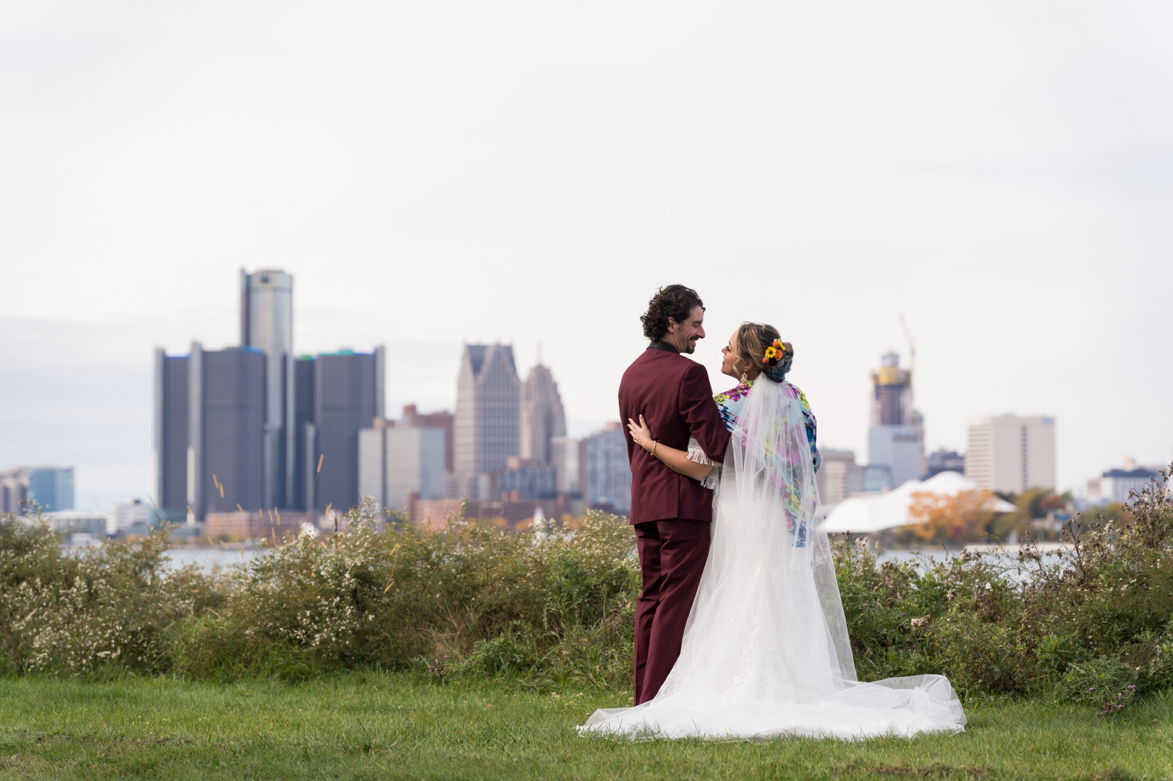 A bride and groom pose with the Detroit skyline in the background.  