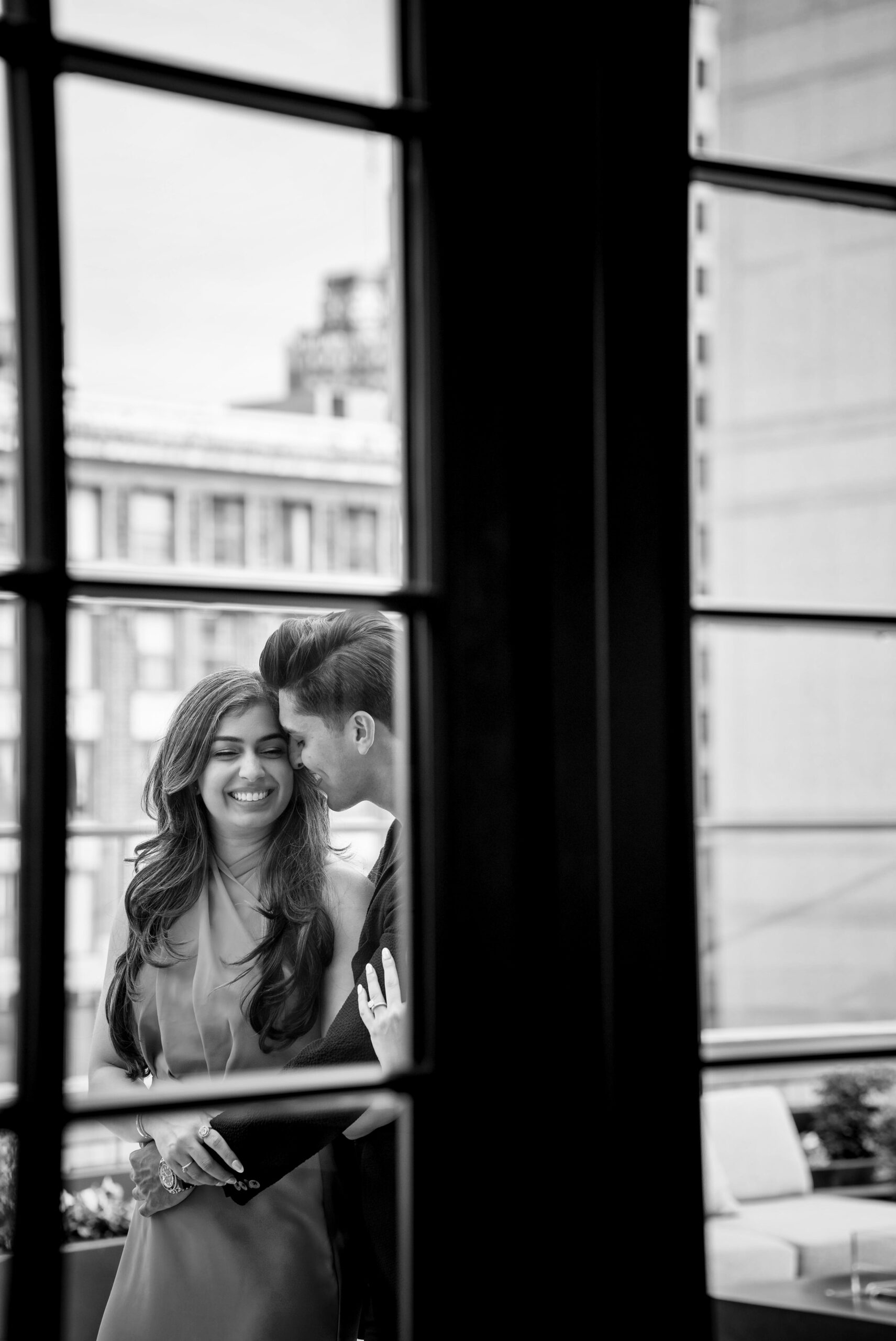 Shot through a window, a couple hugs and laughs during their engagement session at Kamper's rooftop lounge in Detroit.  