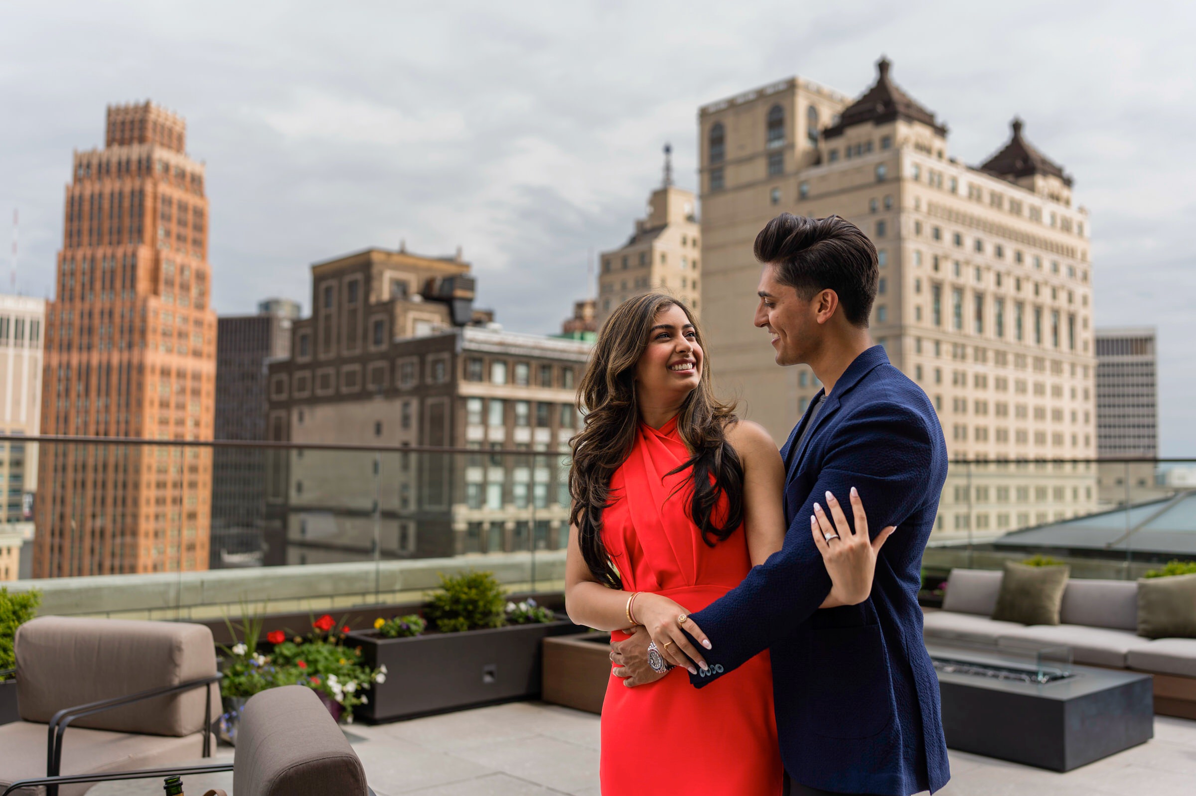 A couple poses during their engagement session at Kamper's rooftop lounge wi the Detroit skyline in the background.  