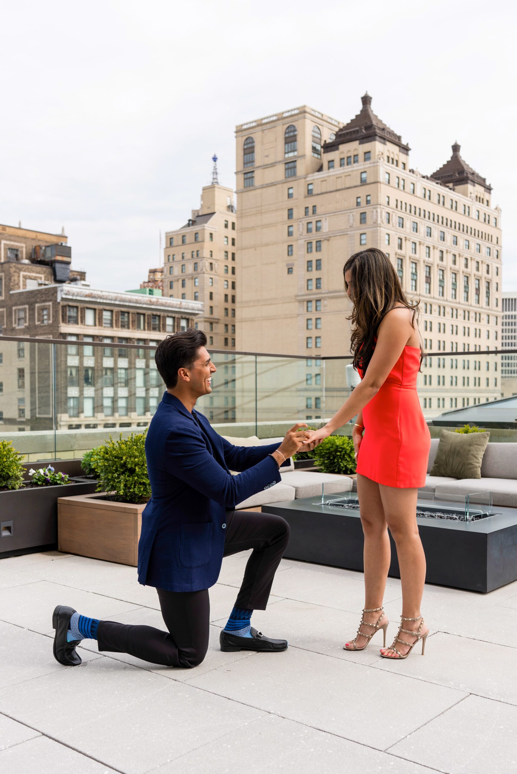 A gentleman takes a knee and places and engagement ring on his fiancé's hand during their Kamper's Rooftop Proposal atop the Book Tower in Detroit.  