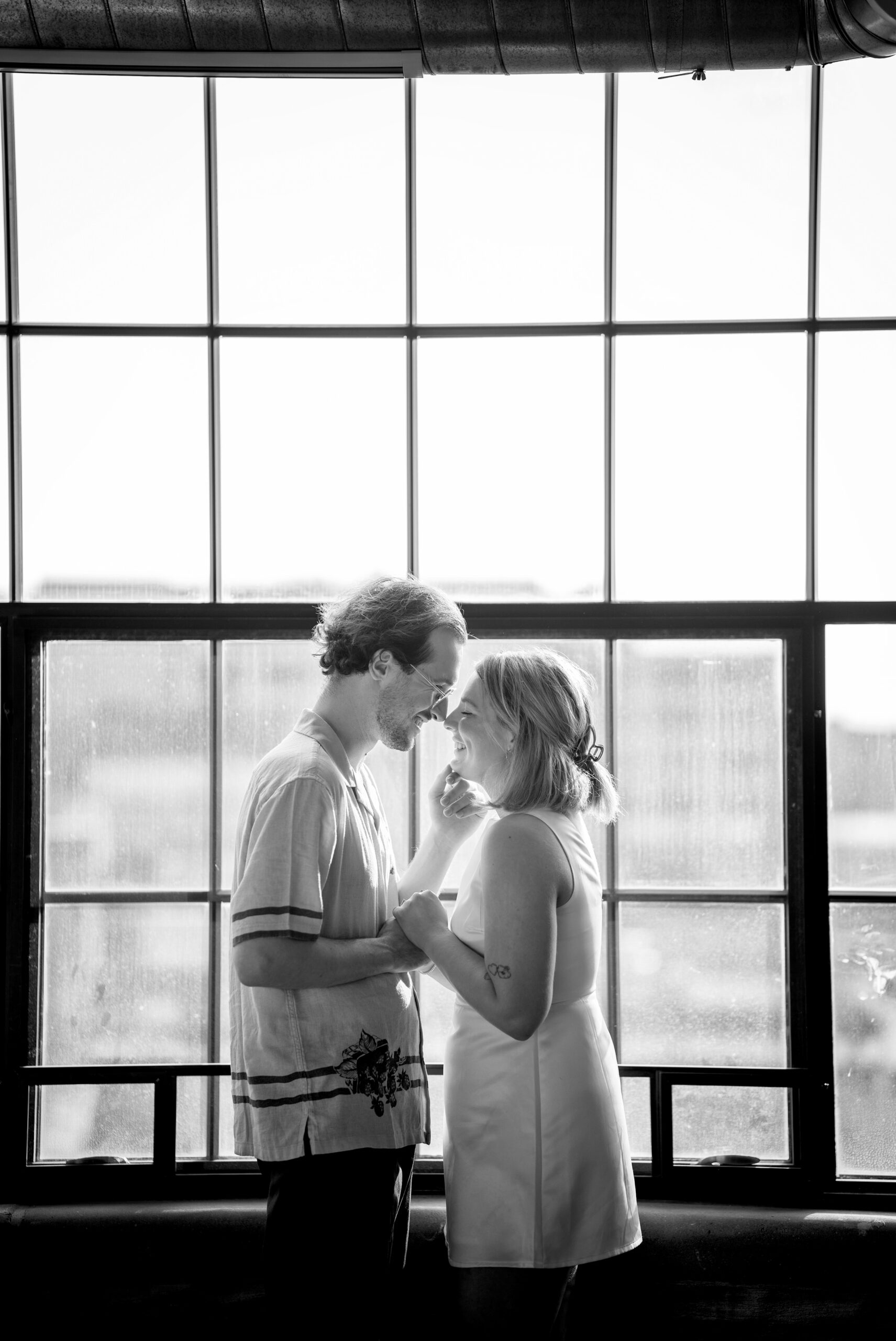 Standing in front of a window, a couple almost kisses during their Detroit lifestyle engagement session.  