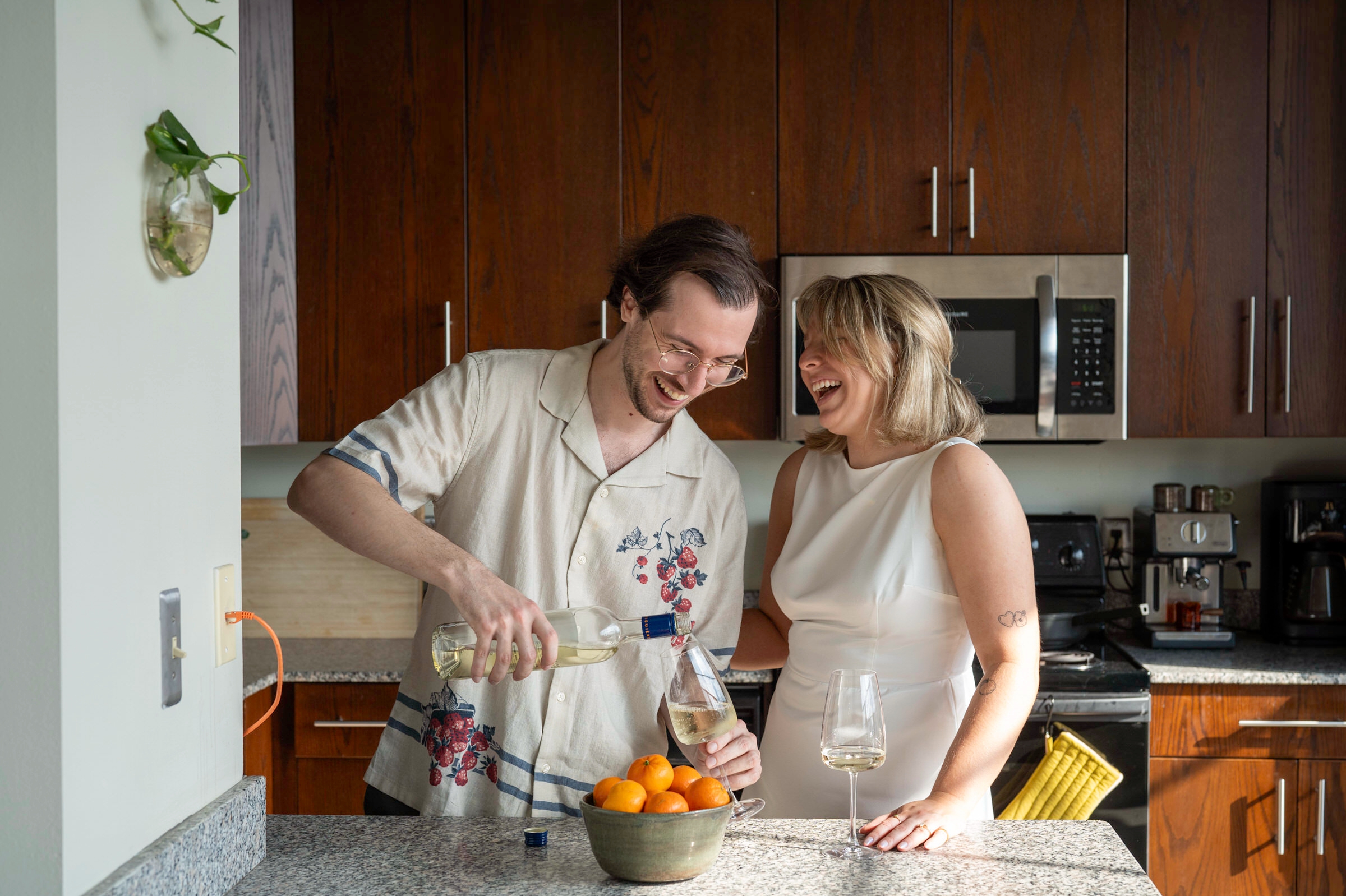 A couple pours a glass of wine  in their kitchen and laughs during their lifestyle engagement session. 