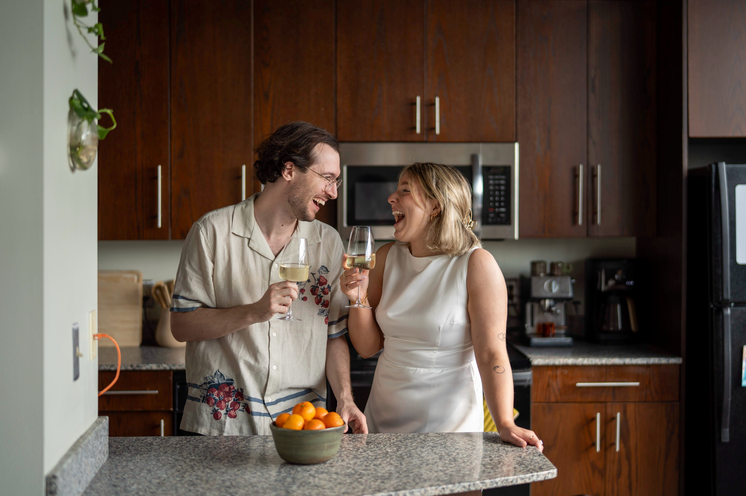 A couple pours a glass of wine  in their kitchen and laughs during their lifestyle engagement session. 