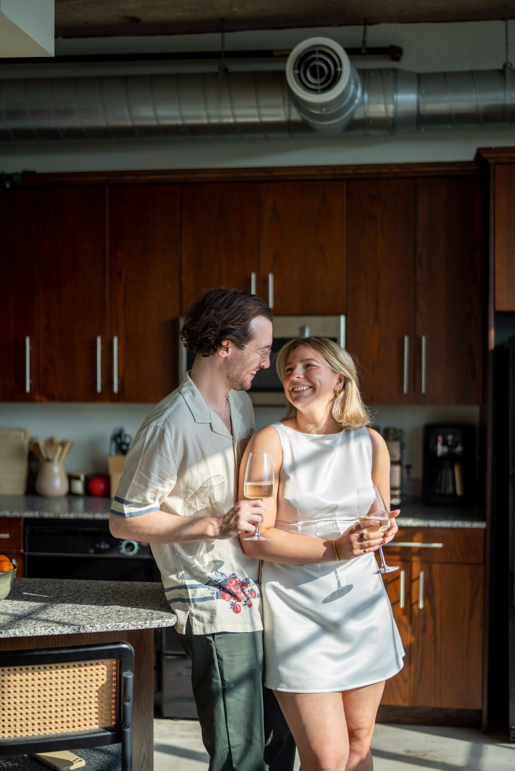 With window frame shadows on them, a couple looks at each other and smiles in their Detroit kitchen during their lifestyle engagement session. 